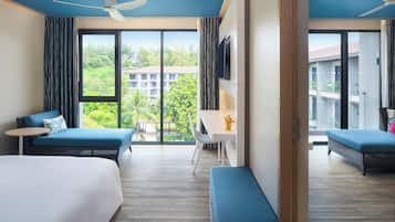 A hotel room with two beds, a desk, and a balcony with a view of buildings and greenery.