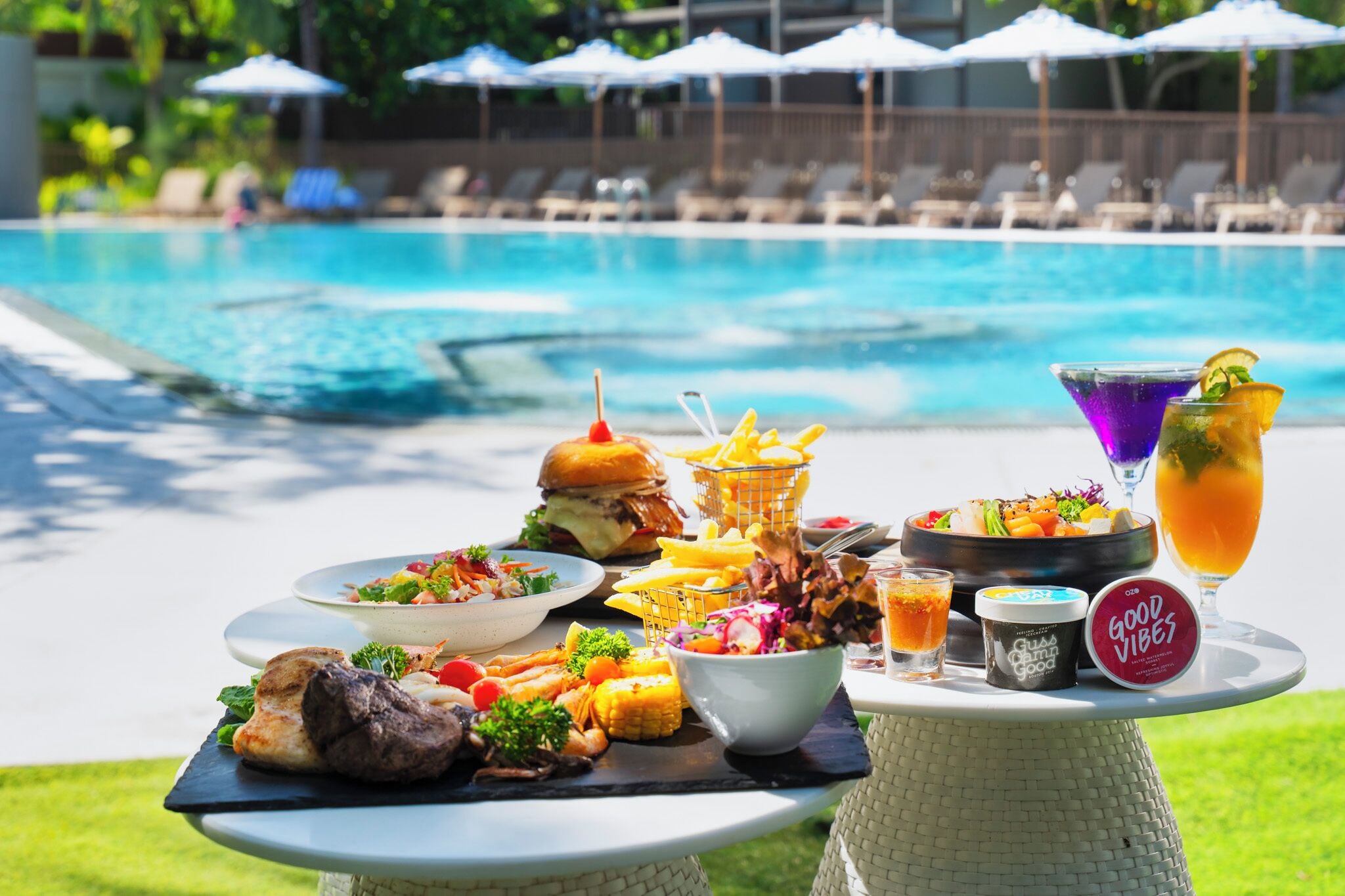 A poolside dining setup with a variety of foods and drinks, including burgers, salads, and a martini.