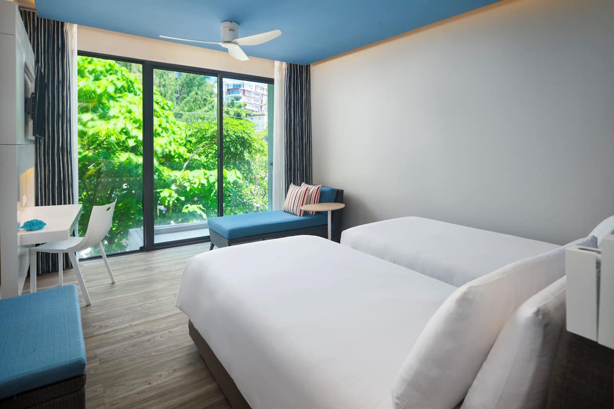 A hotel room with a bed, a desk, a chair, and a view of greenery outside.