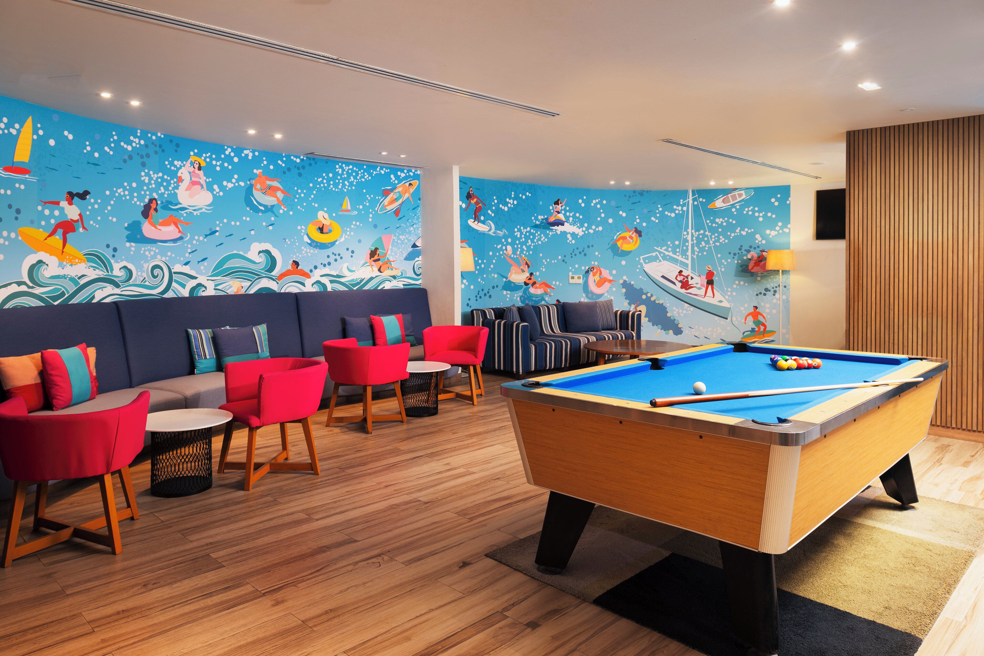 A pool table in a room with a mural of swimmers and boats, red and blue seating, and wooden flooring.