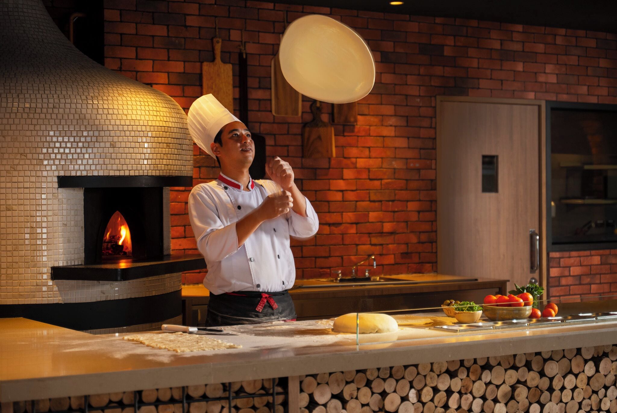 A chef in a kitchen with a brick wall, a wood-fired oven, and a counter with ingredients and utensils.