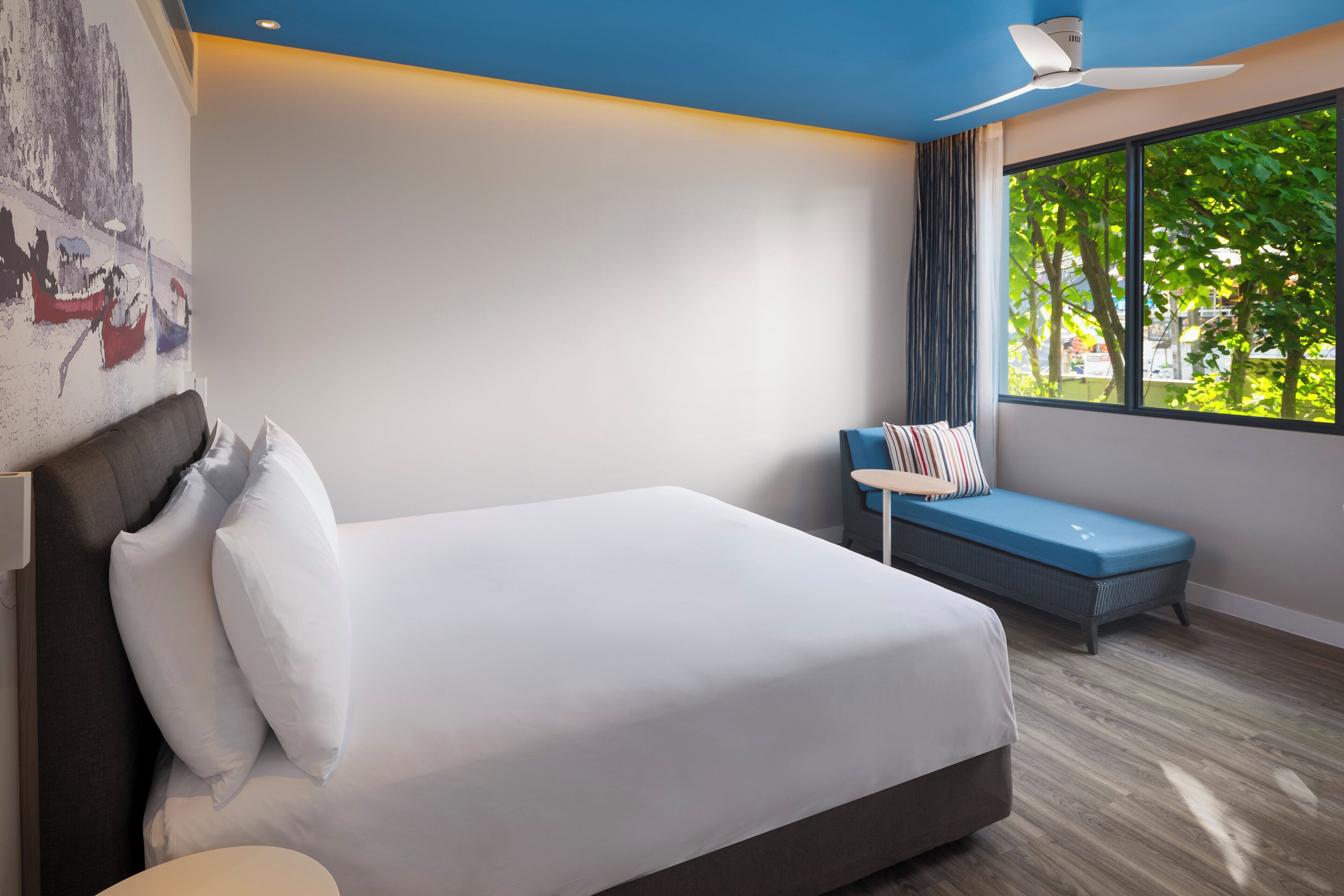 A modern hotel room with a large bed, a small seating area, a ceiling fan, and a large window with a view of trees.