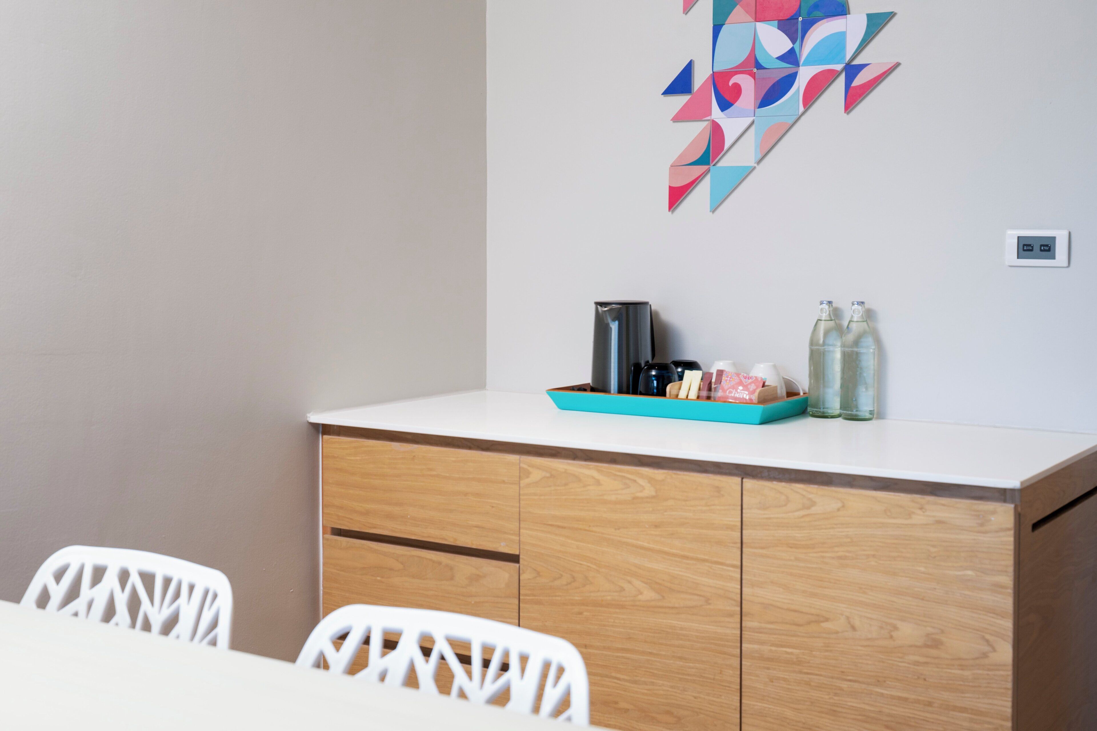 Una habitación moderna con una mesa blanca, sillas blancas, un aparador de madera y una obra de arte geométrica de colores en la pared.