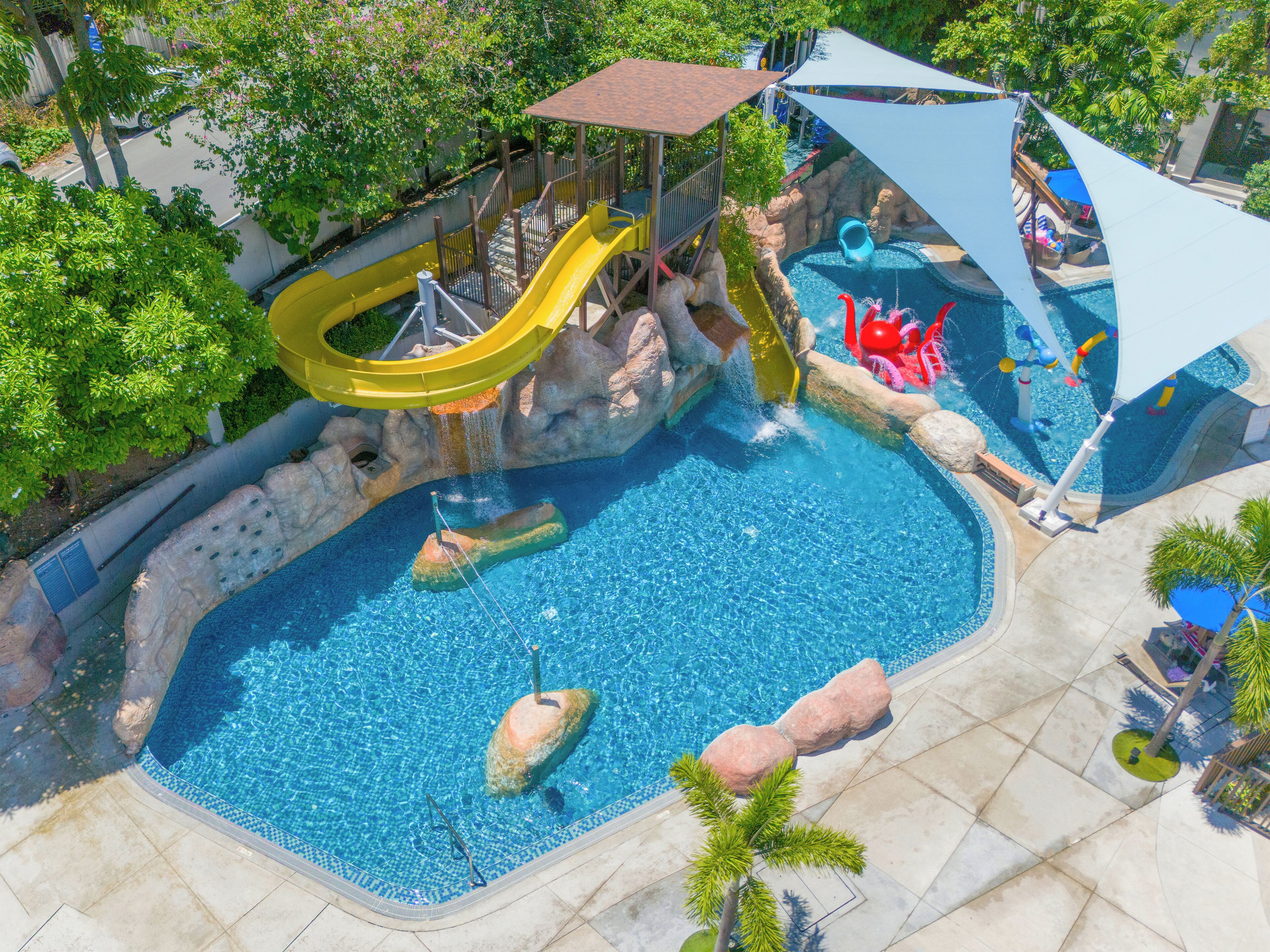 A swimming pool with a water slide, rock formations, and a shaded area with umbrellas.