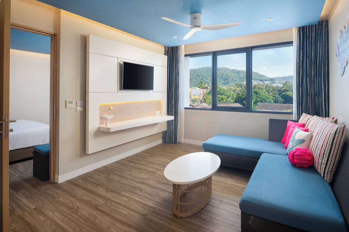 A modern hotel room with a bed, a sofa, a TV mounted on the wall, and a large window offering a view of the outdoors.