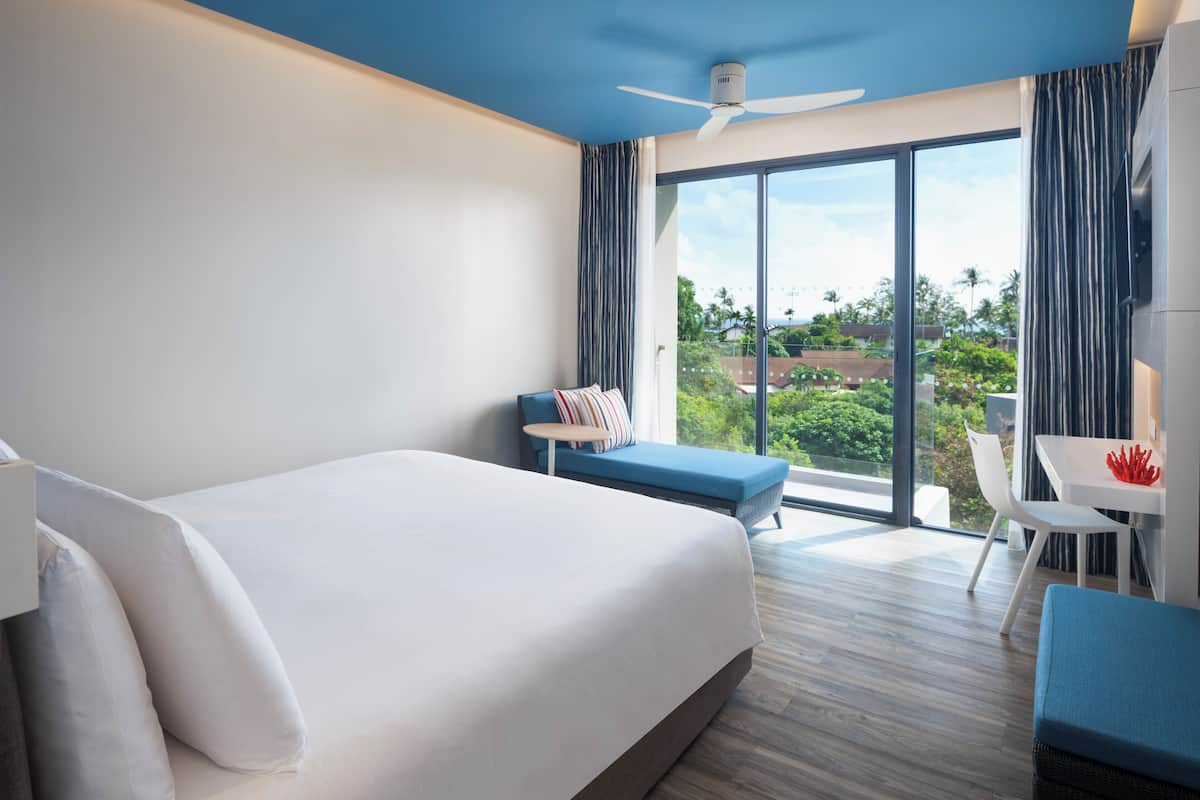 A modern hotel room with a large bed, a small seating area, a dining table, and a balcony with a view of greenery.