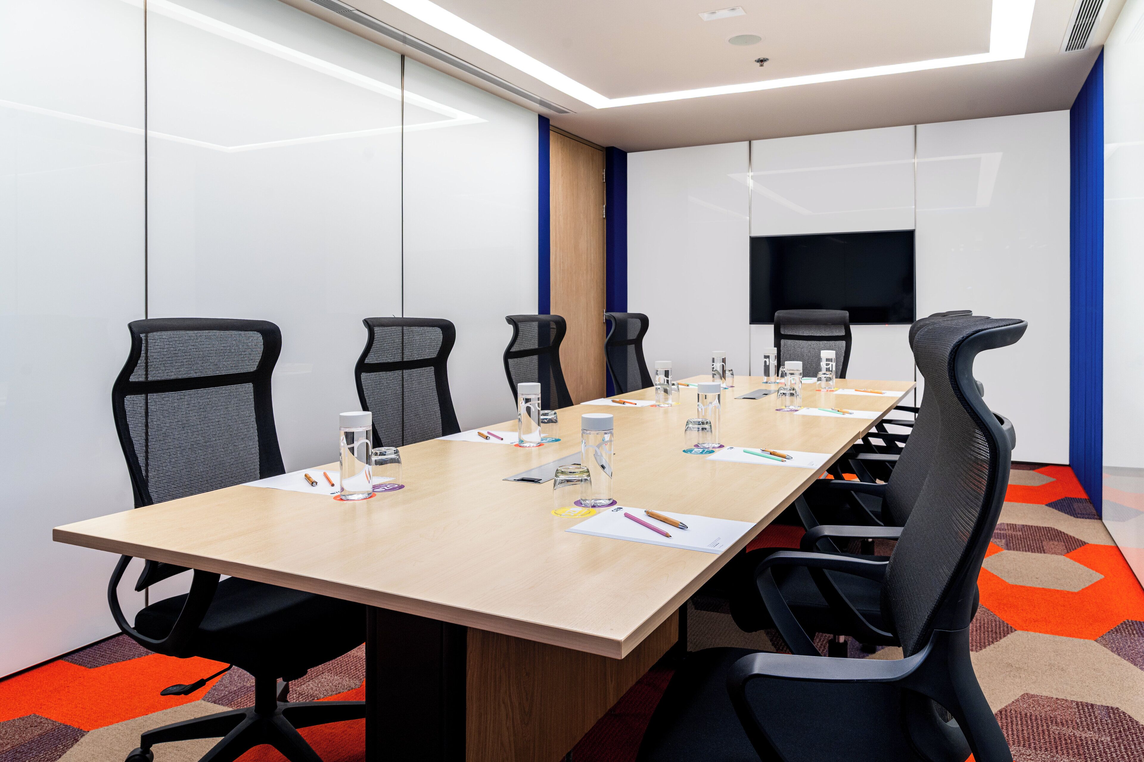 A conference room with a long table, six chairs, and a flat-screen TV on the wall.