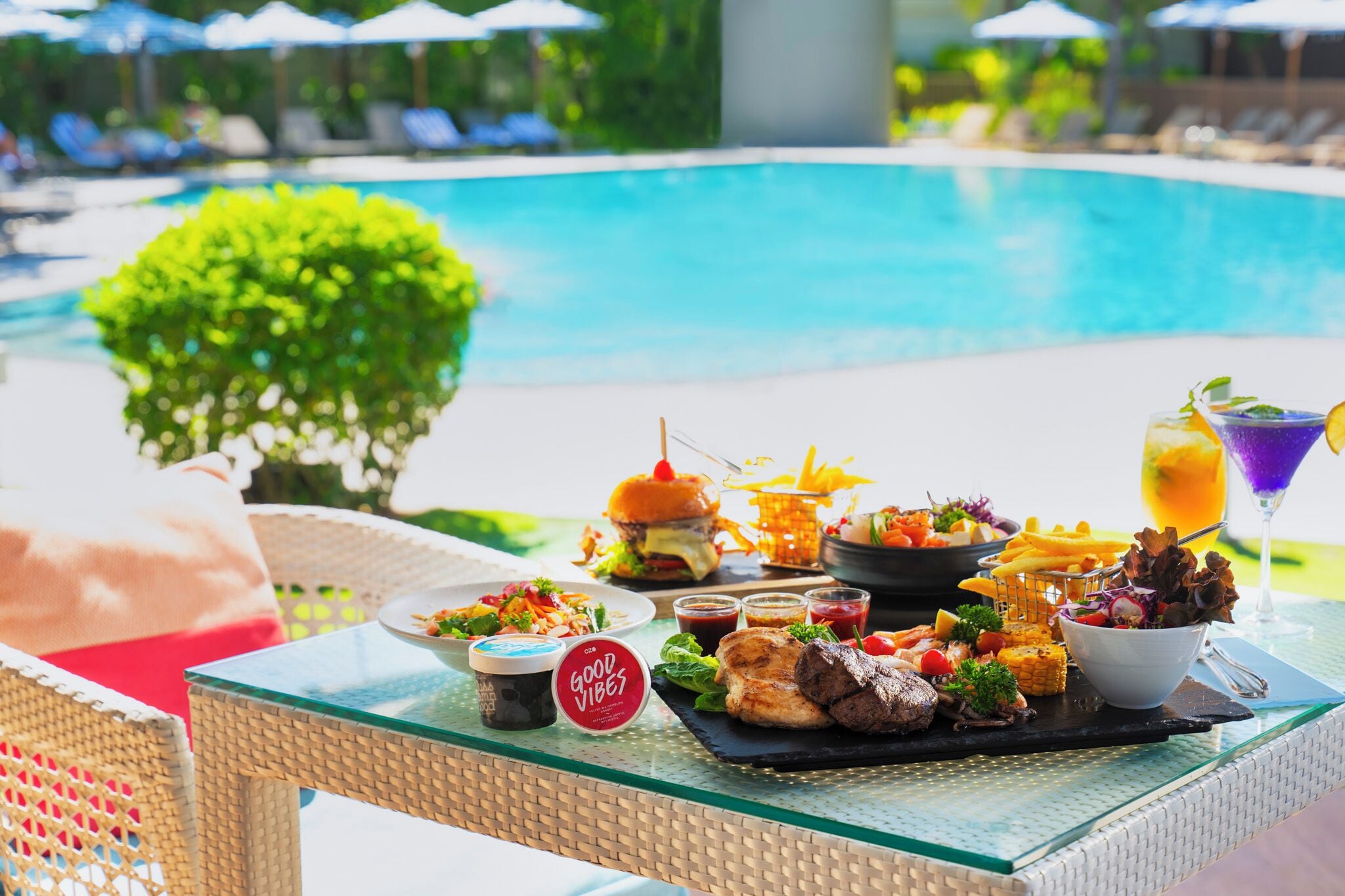 A poolside dining setup with a variety of food and drinks, including burgers, salads, and a martini.