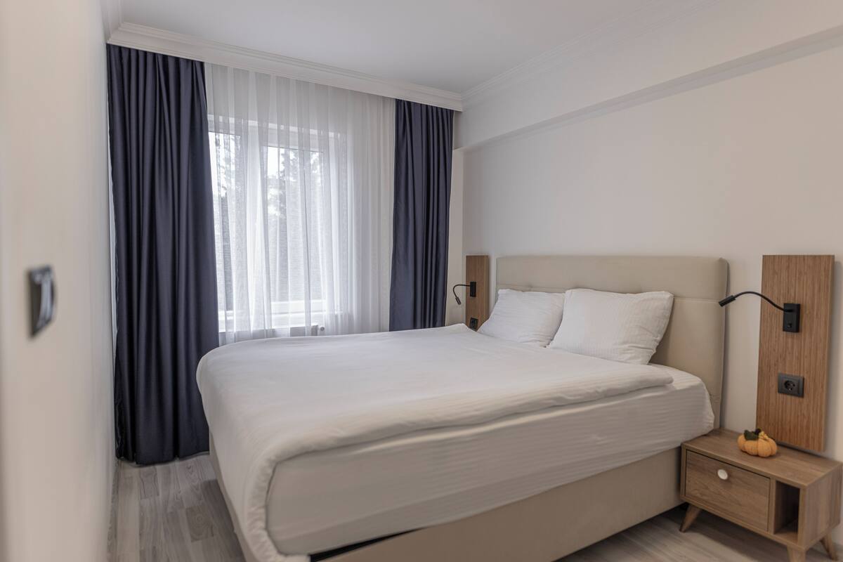 Quarto de hotel com uma cama grande, mesa de cabeceira e janela com cortinas.