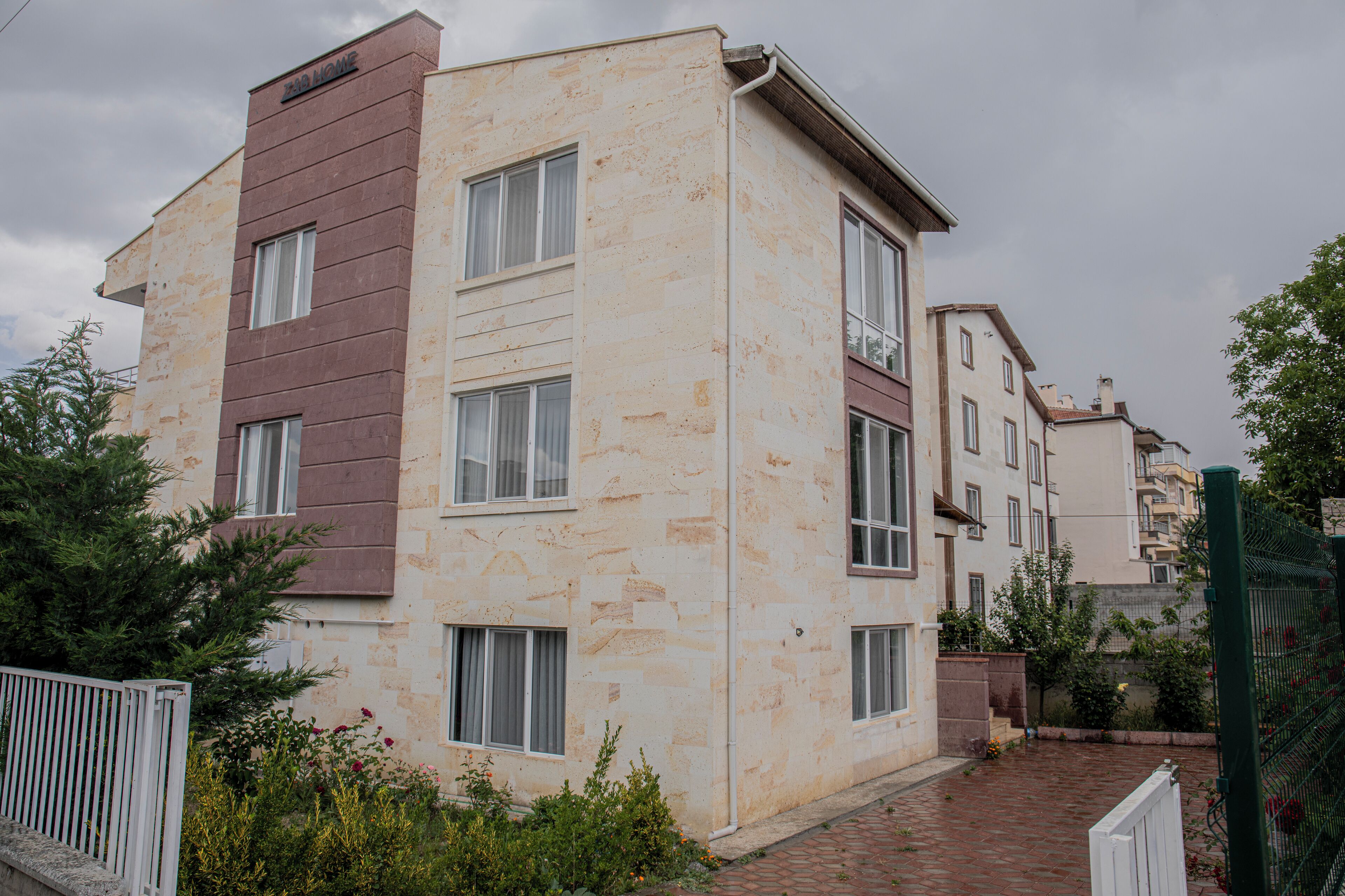 Um edifício residencial de vários andares com paredes externas em tons de bege e castanho, várias janelas e um caminho de tijolos que leva à entrada.