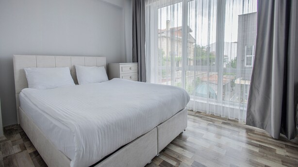 Ein modernes Schlafzimmer mit einem großen Bett, einem Nachttisch und Blick ins Freie durch transparente Vorhänge.