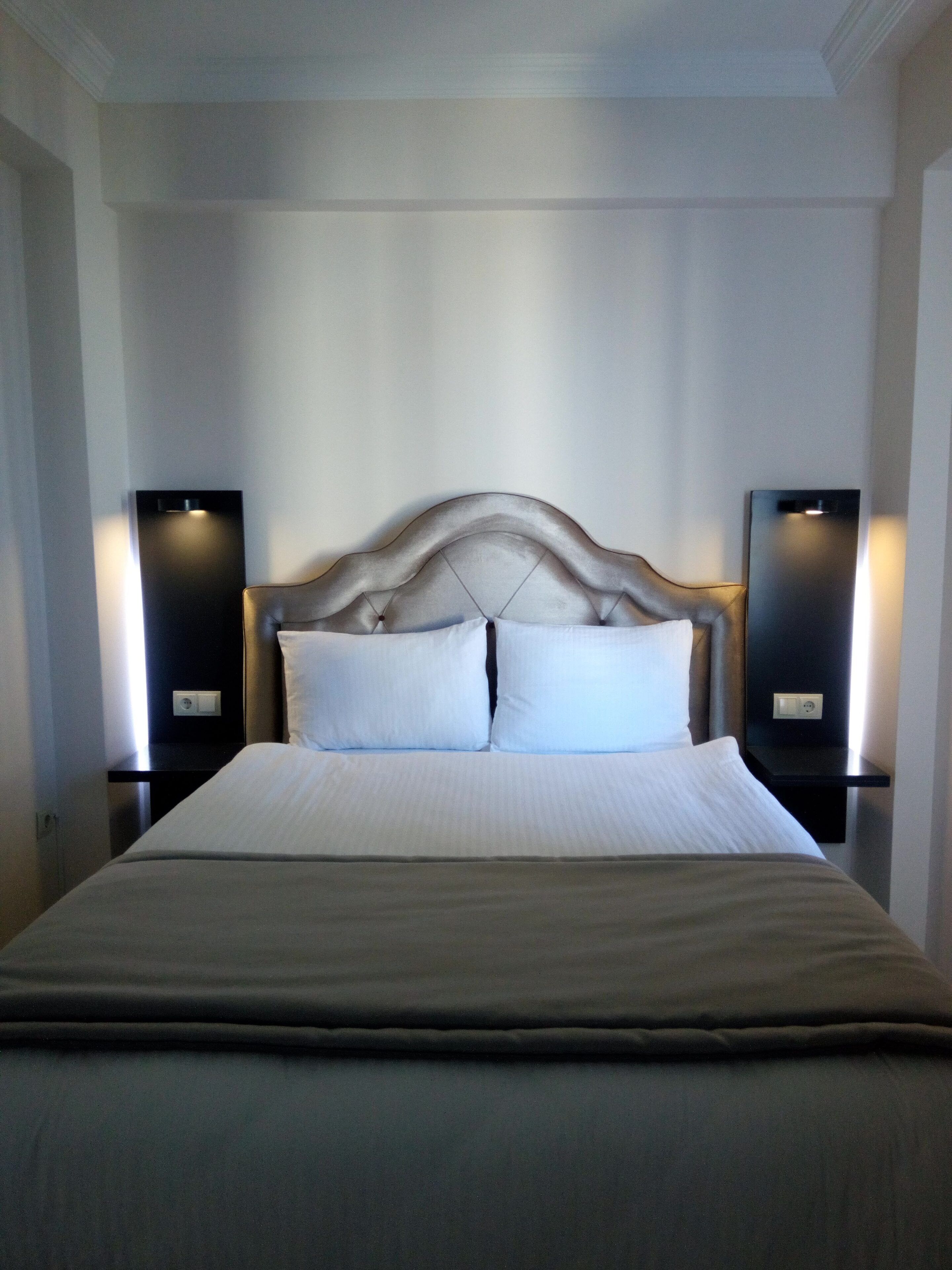 Een netjes opgemaakt bed met een gestuufd hoofdbord, twee nachtkastjes met speakers en een effen muur.