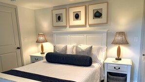 Un dormitorio ordenado con una cama, dos mesitas de noche, una lámpara, una cesta y tres cuadros enmarcados en la pared.