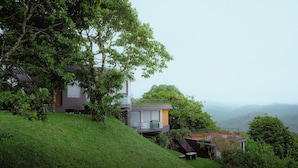 Une maison moderne dotée d’un balcon, entourée d’une végétation luxuriante et avec des collines en arrière-plan.