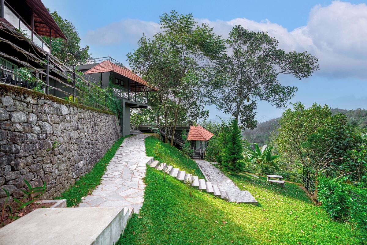 A stone pathway leading to a building with a red-tiled roof, surrounded by lush greenery and trees.