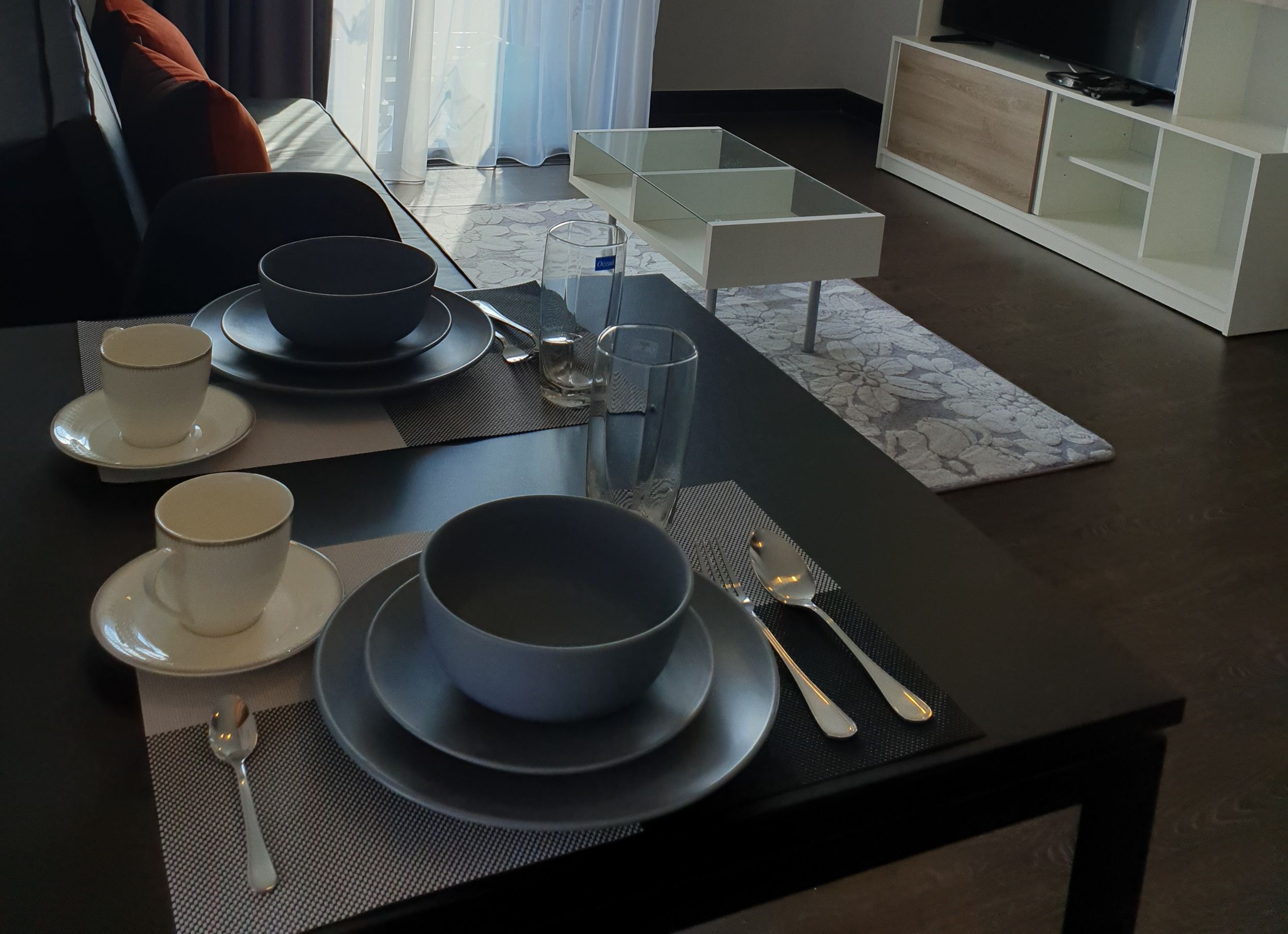 Een eettafel gedekt met borden, kommen, bekers, bestek en glazen, met een modern interieur en een televisie op de achtergrond.