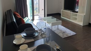 En matplats med ett bord för fyra personer, en soffa med kuddar, en tv på ett tv-bord och utsikt över omgivningen genom en skjutdörr.