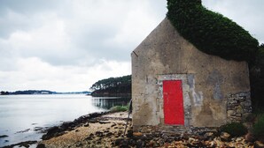 Un petit bâtiment en pierre avec une porte rouge et un toit vert, situé au bord d’une étendue d’eau calme.