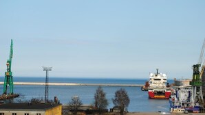 Um porto com um grande navio de carga atracado, guindastes e edifícios industriais.