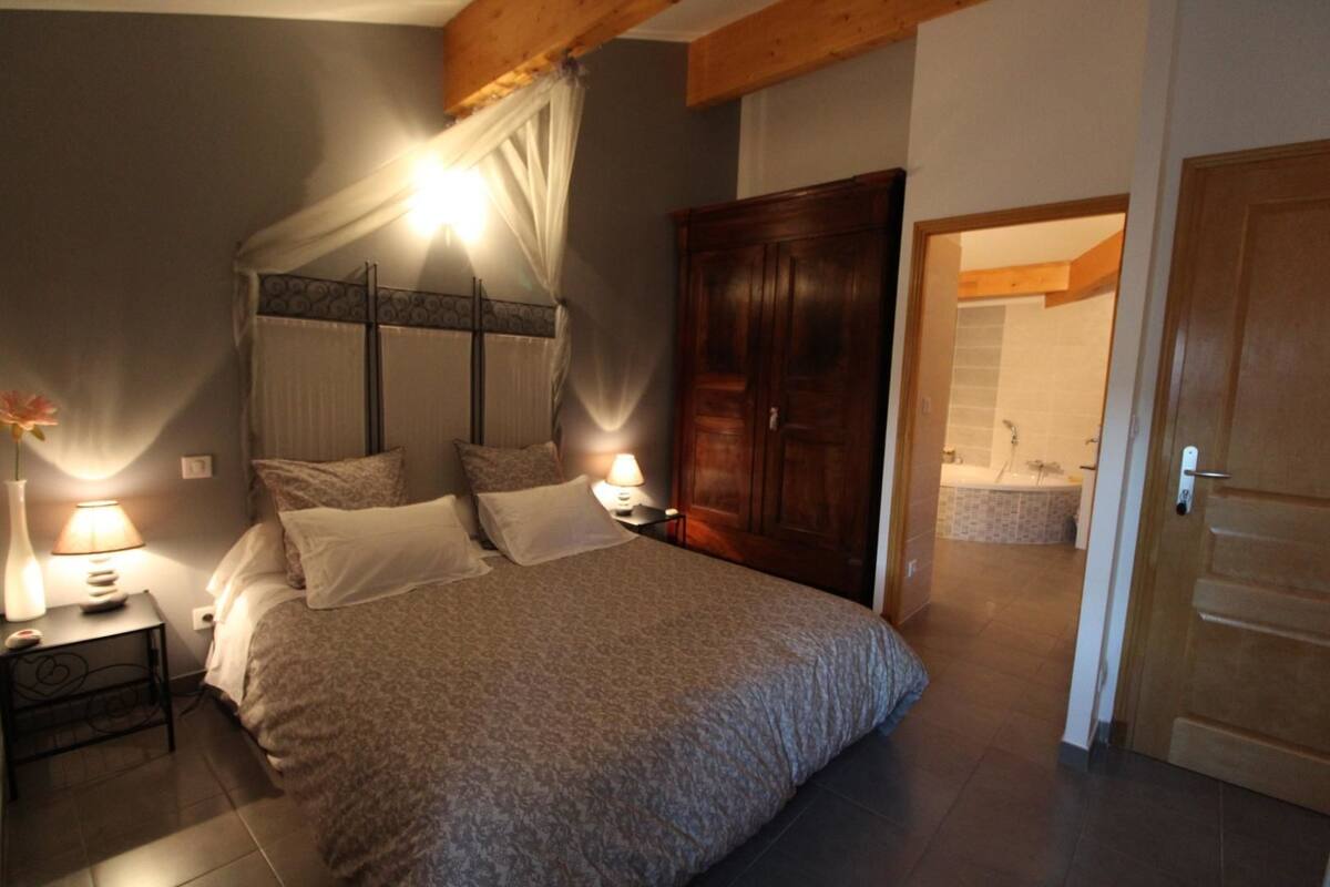 A bedroom with a large bed, a wooden wardrobe, a nightstand with a lamp, and a bathroom visible through an open door.