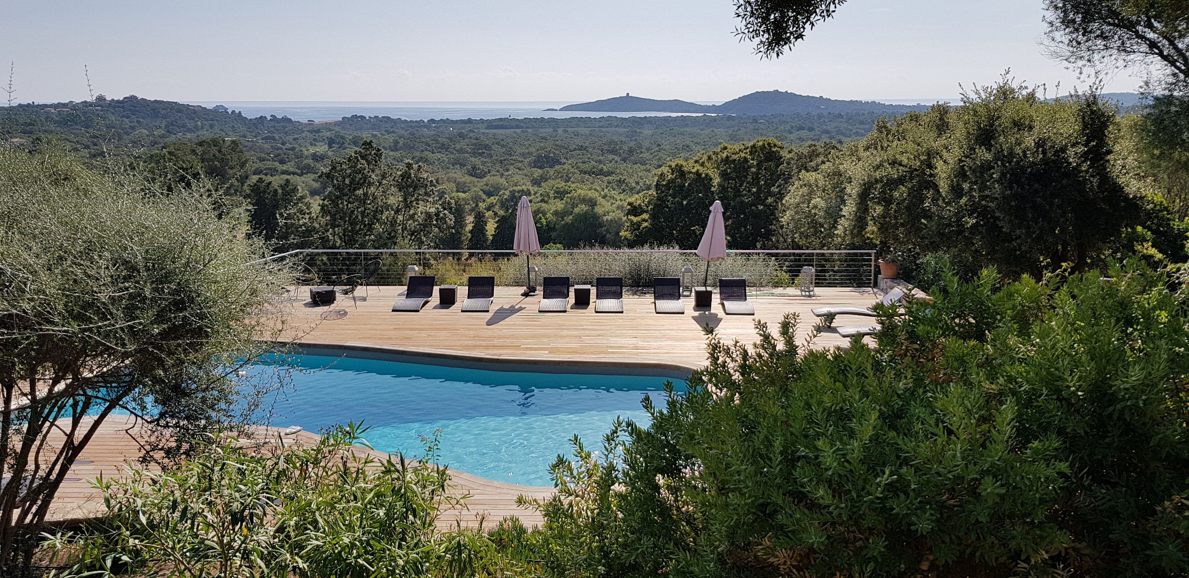 A poolside area with lounge chairs and umbrellas, surrounded by greenery and overlooking a scenic landscape.
