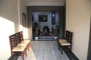 A hallway with a marble floor leading to a seating area with wooden chairs and a small table. There are potted plants and a clock on the wall. - Vandana Residency (Varanasi)