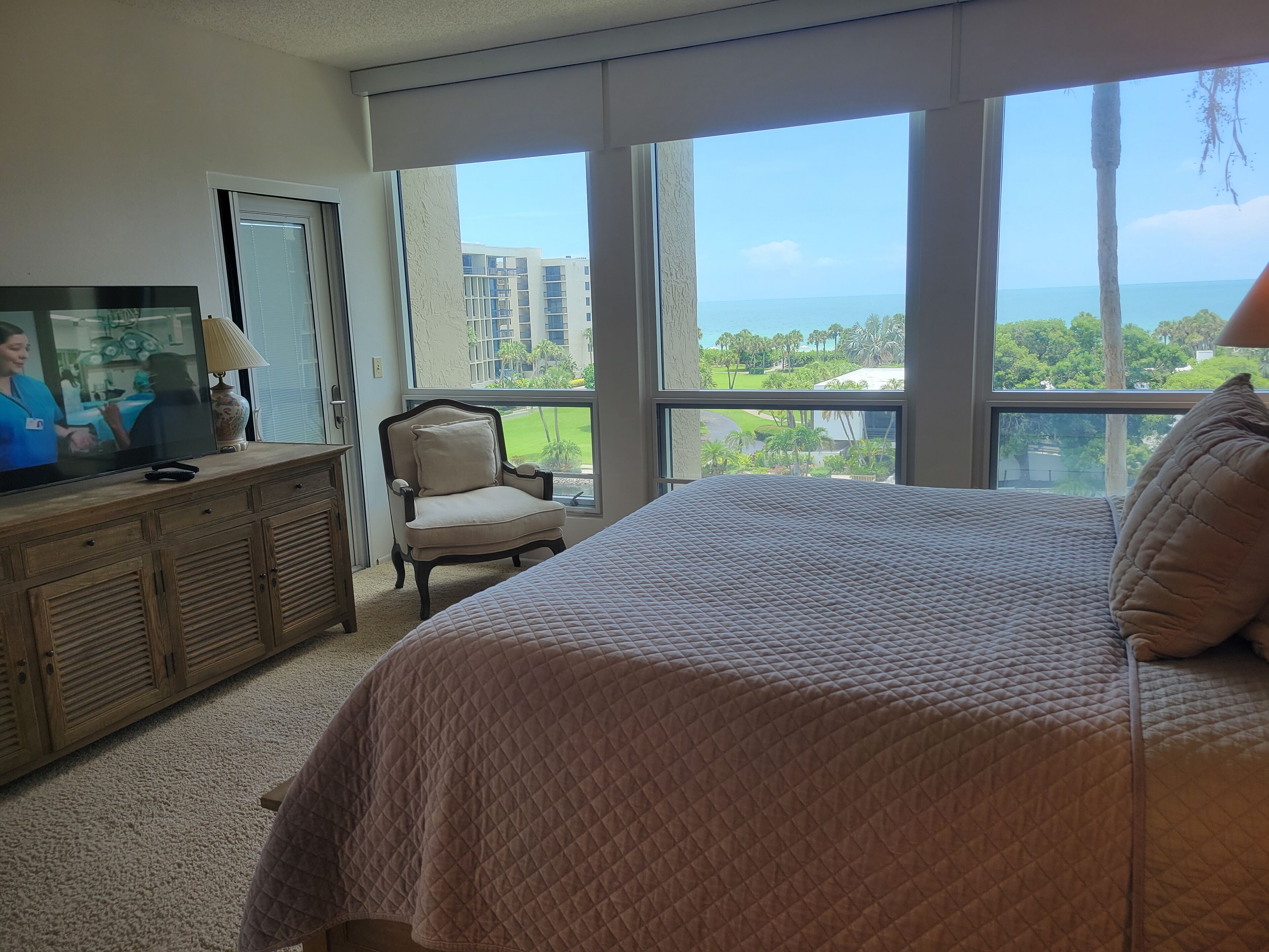 A bedroom with a large bed, a television on a wooden dresser, a chair, and a view of buildings and greenery through large windows.