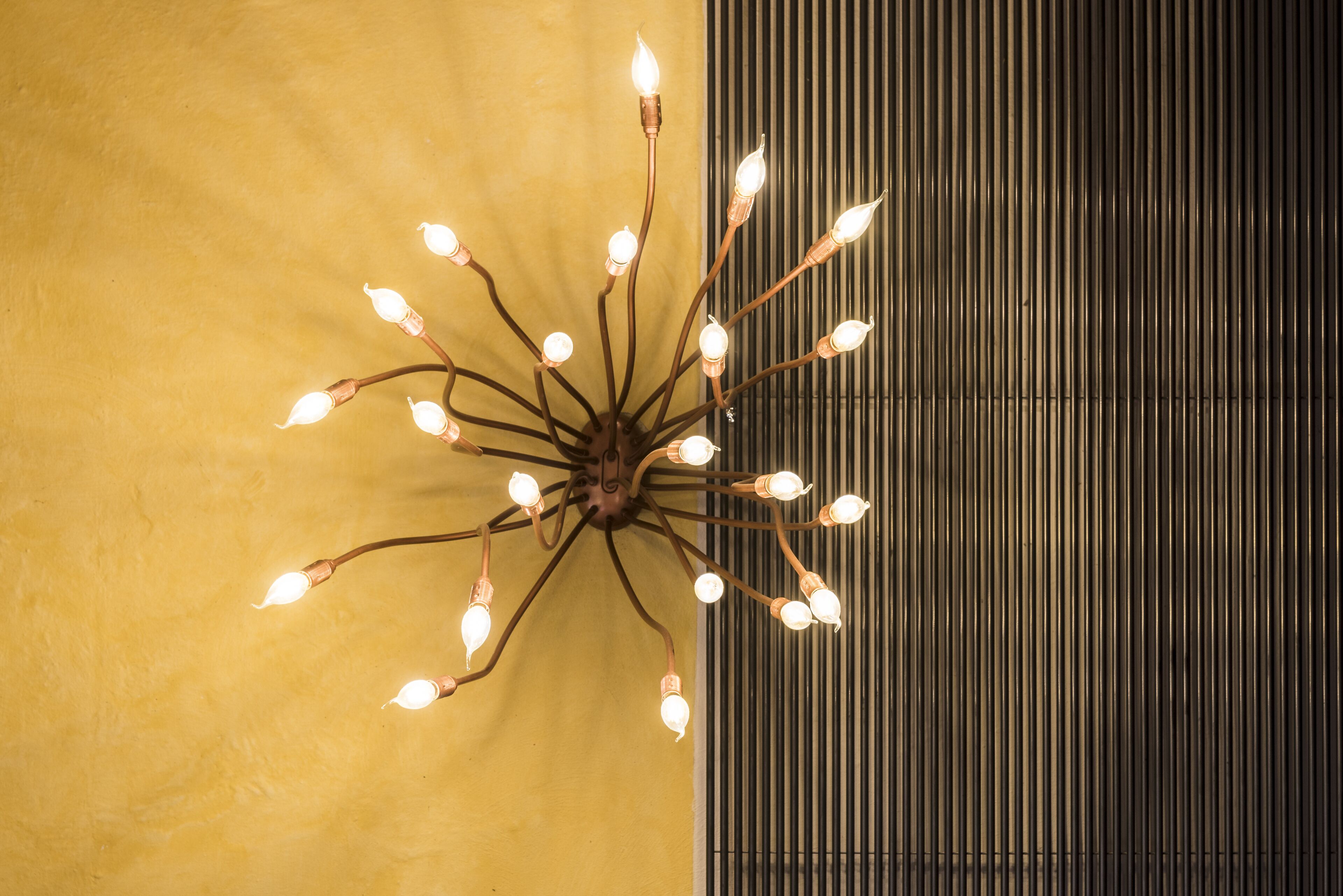 Un lustre doté de plusieurs ampoules, placé devant un mur jaune et un panneau métallique vertical.