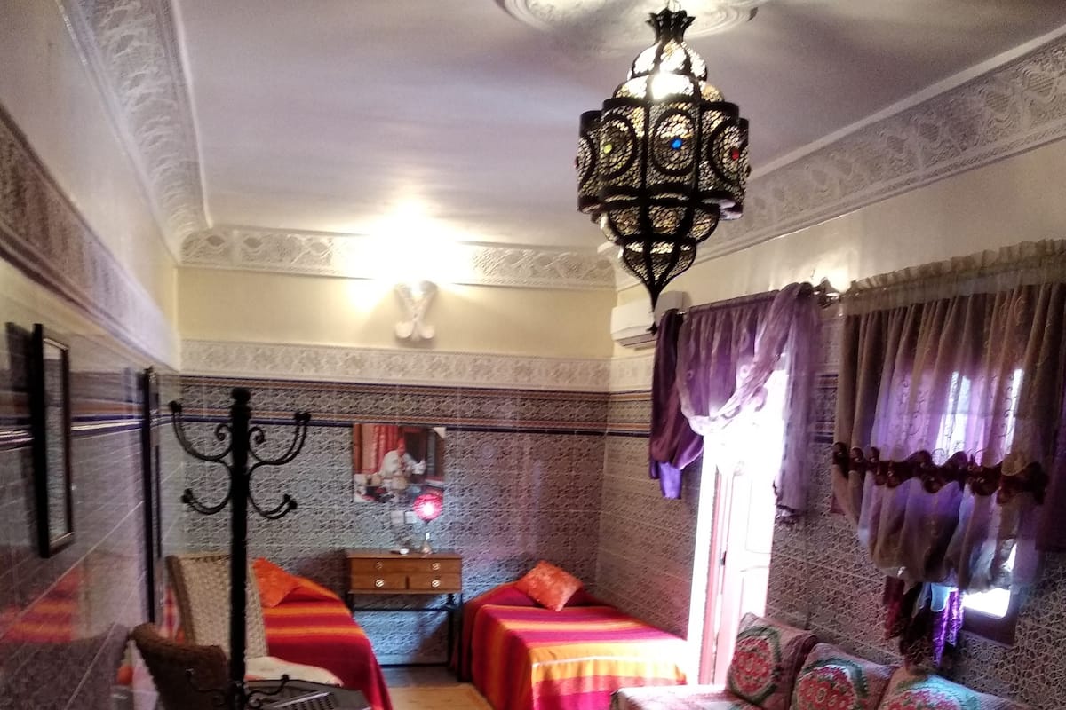 A traditional Moroccan room with a patterned floor, a bed with striped bedding, a sofa with cushions, a wooden dresser, and a hanging lamp.
