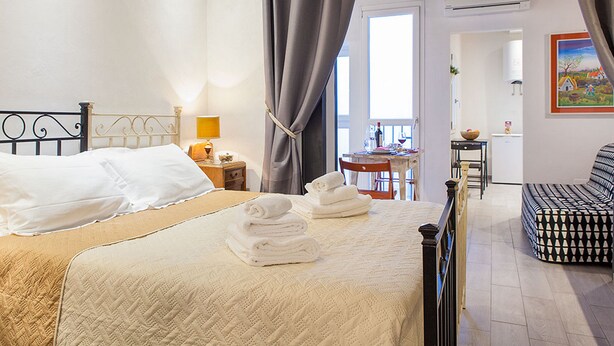 A bedroom with a bed, a bedside table, a lamp, a chair, and a view of the dining area. - Santa Croce Apartment (Florence)