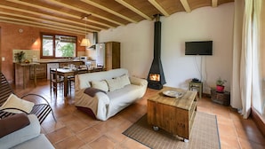 Un salon chaleureux avec un canapé, une table basse et une cheminée. Au fond, on aperçoit une cuisine avec une table à manger et des chaises.