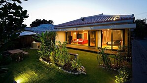 Une maison moderne avec un toit en tuiles, un salon avec de grandes baies vitrées et des meubles rouges, et un jardin bien entretenu.