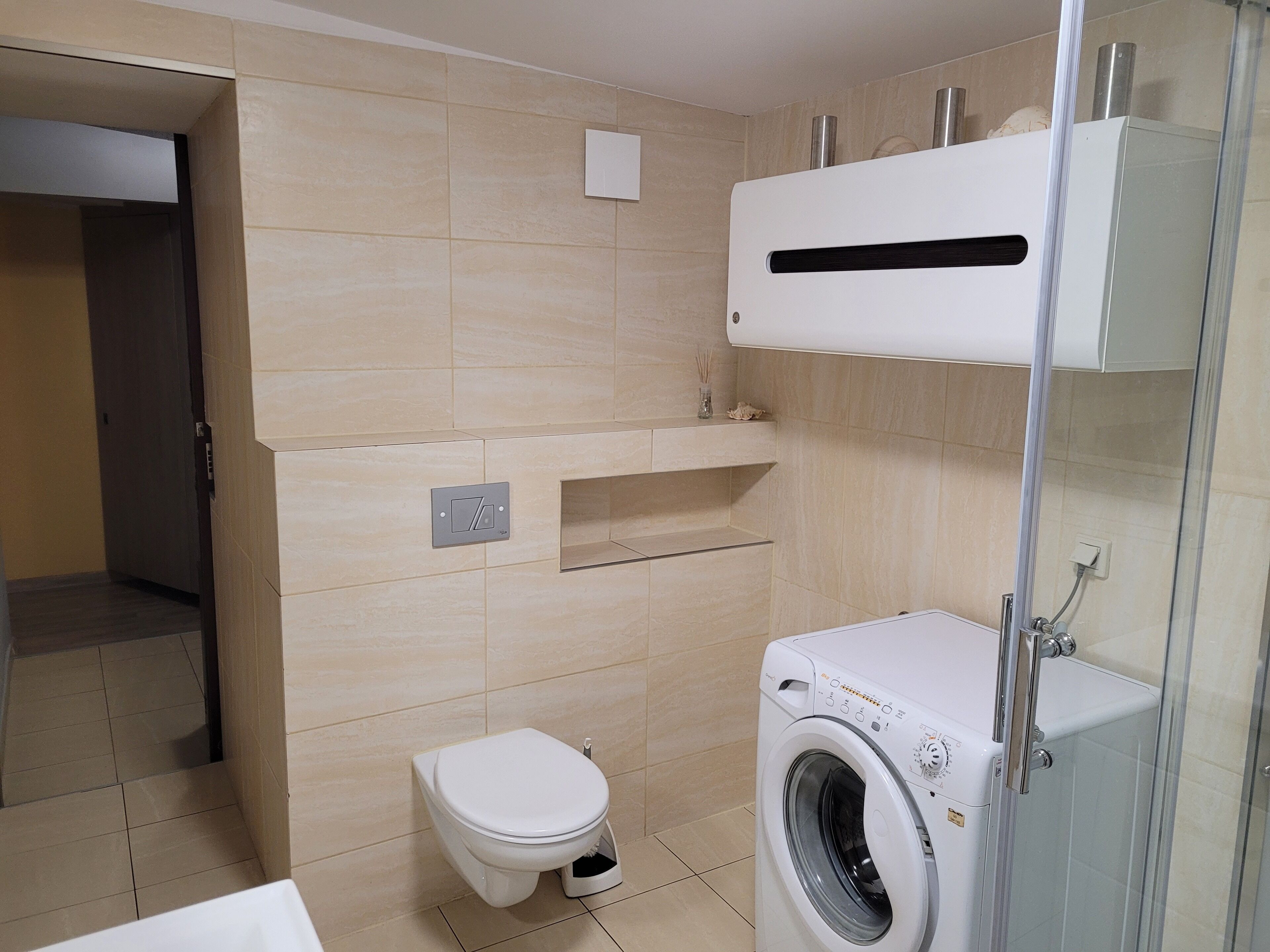 Casa de banho moderna com sanita, máquina de lavar roupa e cabine de duche em vidro.