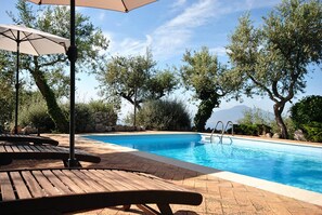 Une piscine entourée d’arbres et d’un mur de pierre, avec des chaises longues et des parasols à proximité.