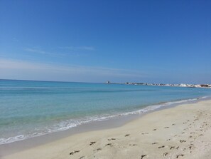 A beach with clear blue water, sandy shore, and buildings in the distance. - Holiday House '' Lu Barocco '' (San Pancrazio Salentino)