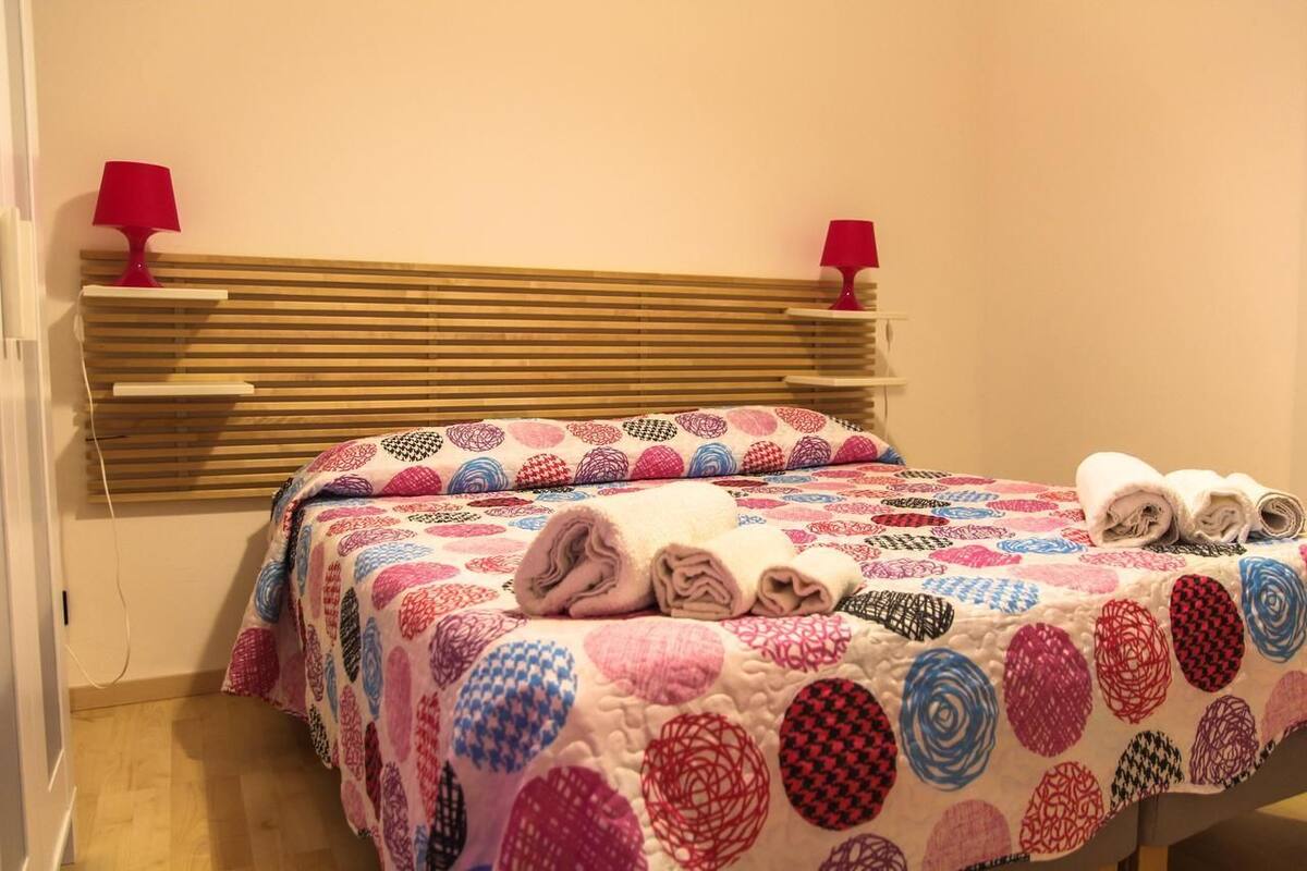 A bed with a colorful quilt, two red lamps on a bedside table, and rolled towels on the bed.