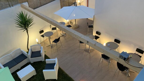 An outdoor patio area with white wicker furniture, including sofas and chairs, arranged around several round tables. There is a palm tree and a wooden pergola structure.