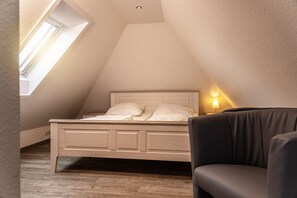 Ein Schlafzimmer mit schrägen Wänden, einem Bett mit weißer Bettwäsche, einer Nachttischlampe und einem dunkel gefärbten Sessel.