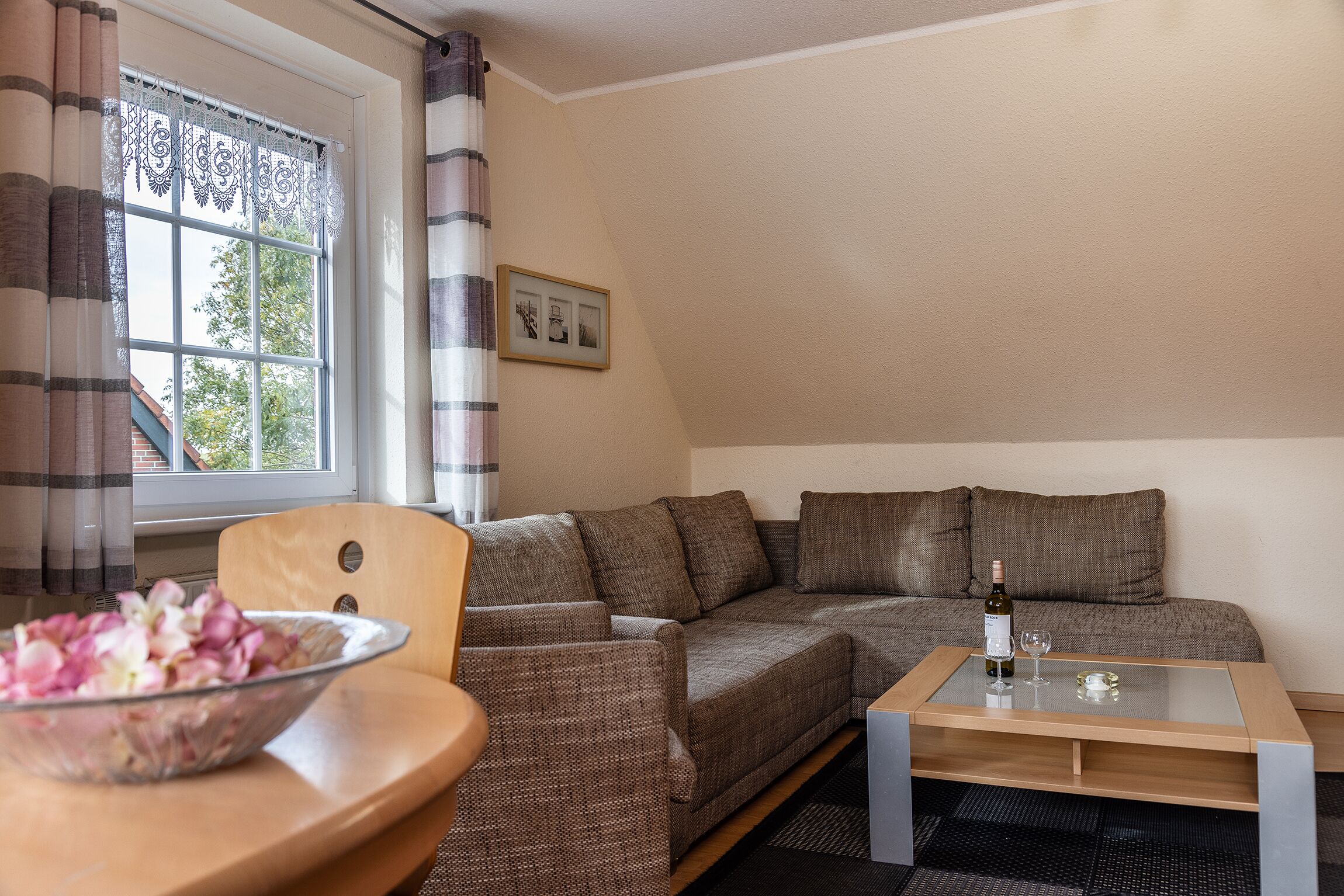 A living room with a sofa, a glass coffee table, a bowl of flowers, and a bottle of wine.