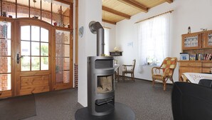 A cozy living room with a wood-burning stove, a wicker chair, a wooden cabinet with books, and a view through a glass door. - Gästehaus, Das Fischerhus (Esens)
