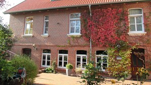A two-story brick building with multiple windows, a covered porch, and a garden area with plants and a hose reel. - Gästehaus, Das Fischerhus (Esens)