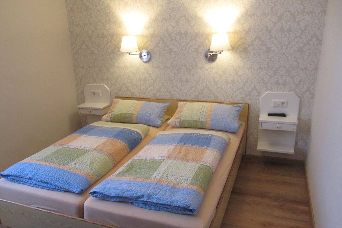 Uma cama de casal com colcha, duas mesas de cabeceira, um criado-mudo e luminárias fixadas na parede.
