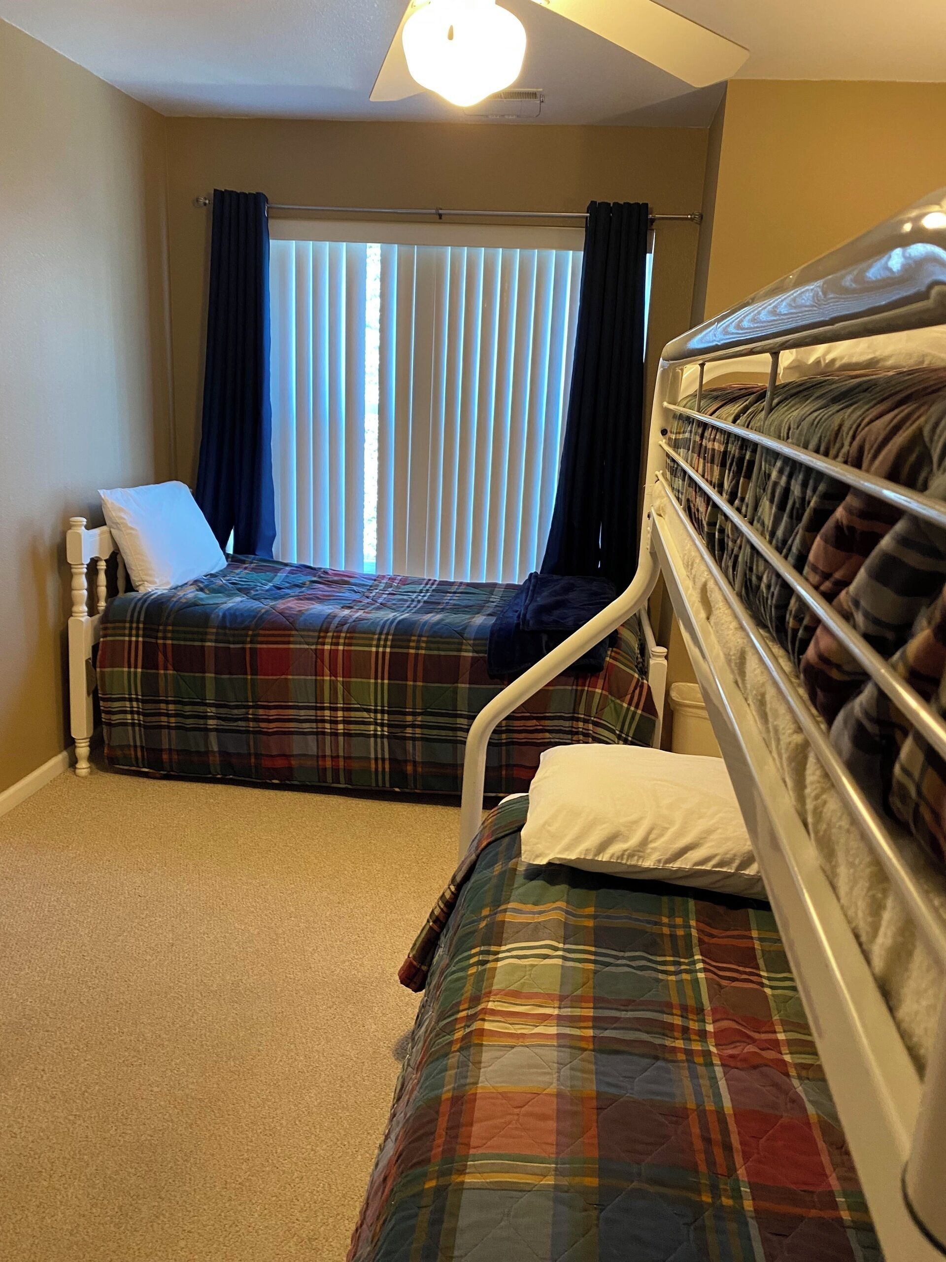 Une chambre avec deux lits, une fenêtre avec des rideaux, un ventilateur de plafond et un sol recouvert de moquette.