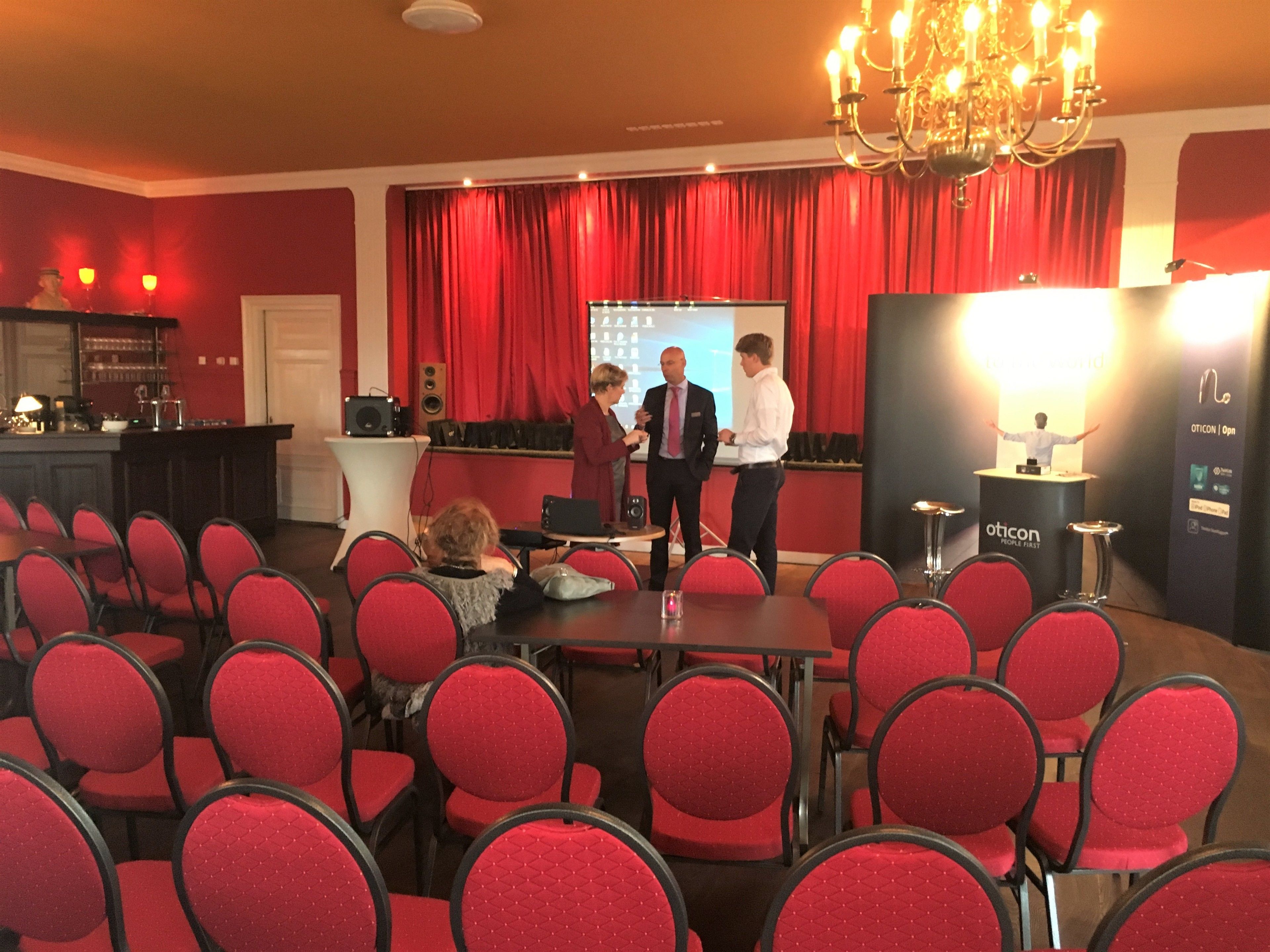 Un salón de conferencias con sillas rojas, un escenario con un atril y una araña de cristal.
