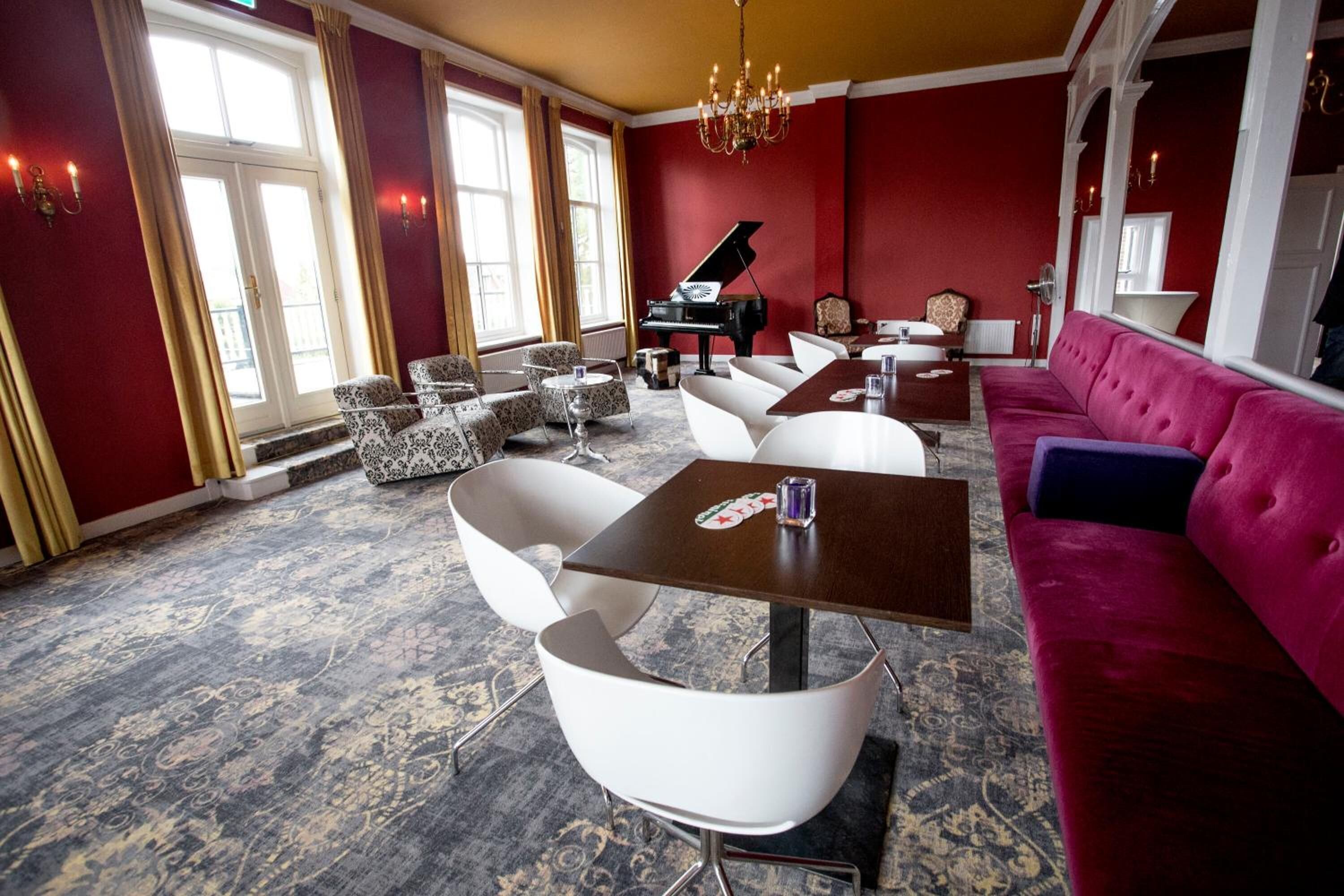 Een kamer met een vleugelpiano, rode muren, een kroonluchter en een luxe rode bank.