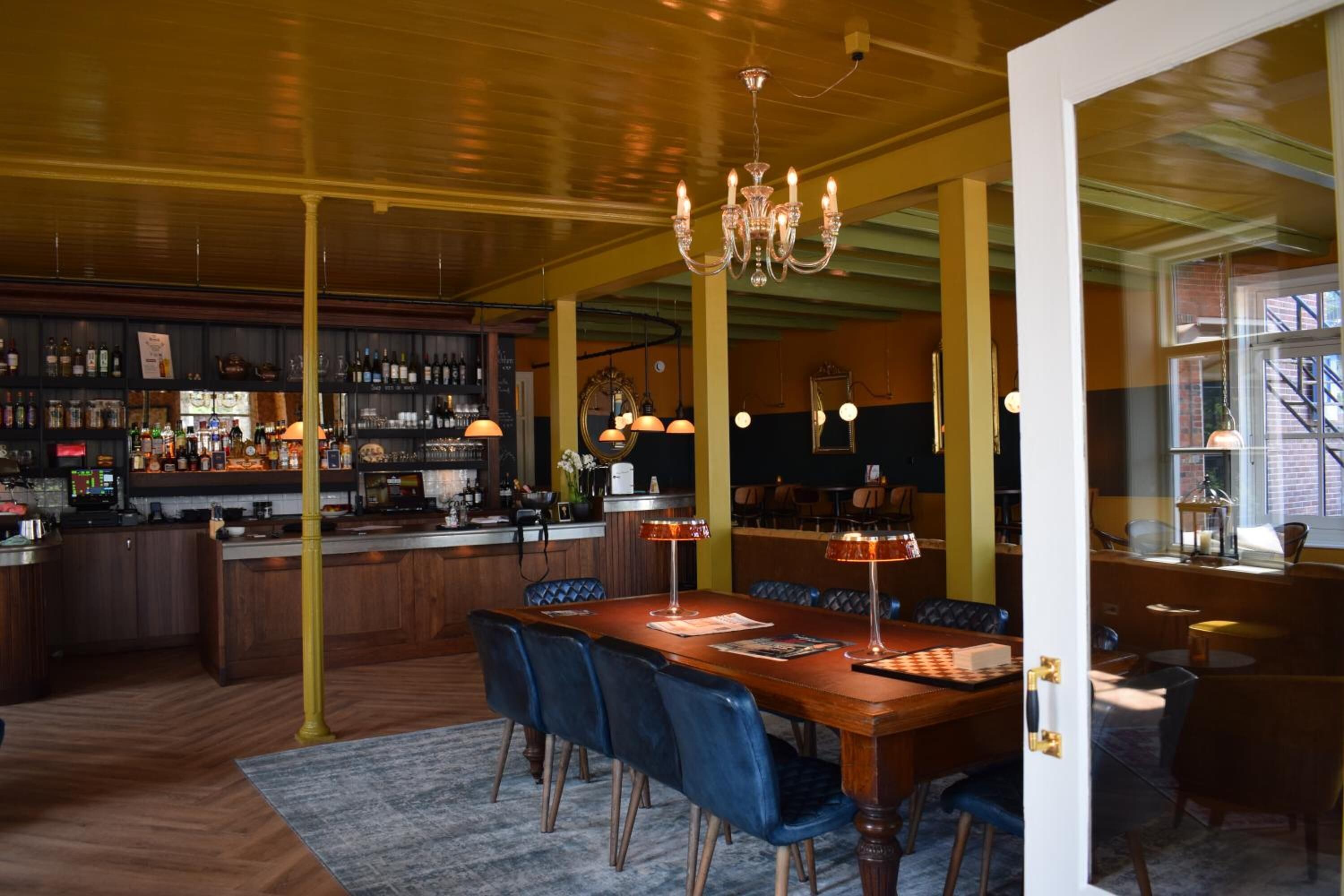 Una zona de bar con mostrador de madera, estanterías con botellas y un espacio de asientos con una mesa de madera y sillas azules.