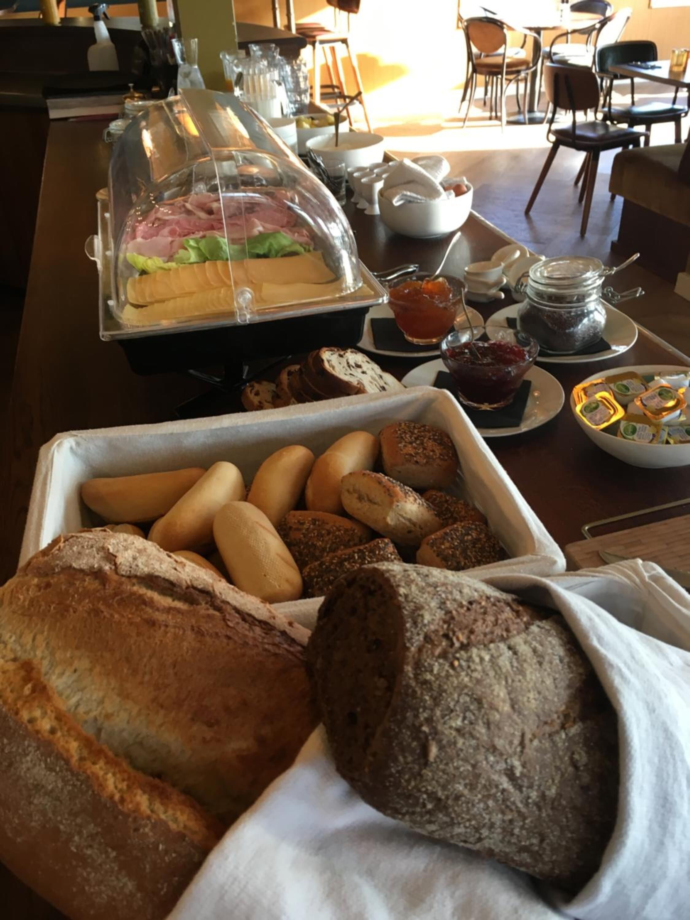 A buffet with various breads and pastries, a glass container with sandwiches, and a jar of jam.