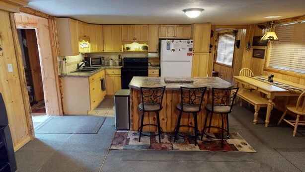 A compact kitchen with wooden cabinets, a refrigerator, a microwave, and a stove. There are three bar stools at the counter. - Yellowstone NP | Sleeps 8 | Fire Pit & BBQ (Island Park)
