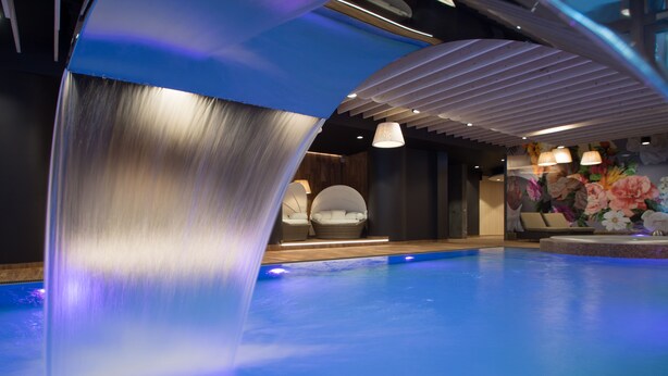 A modern indoor pool area with a waterfall feature, seating by the pool, and a floral mural on the wall.