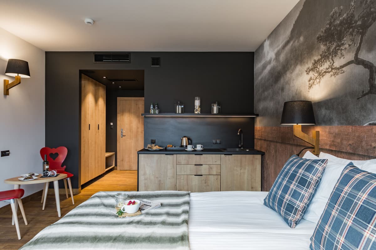 A modern hotel room with a bed, a desk, a chair, a small table, and a kitchen area.